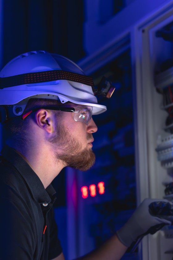 A male electrician works in a switchboard in overalls against the backdrop of emergency lighting. Surveillance continue de votre infrastructure